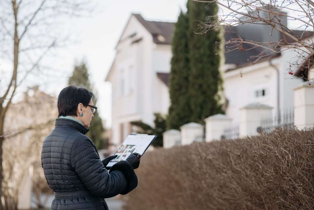 Woman wearing a black winter jacket and glasses examining property documents on a clipboard outside a large suburban house