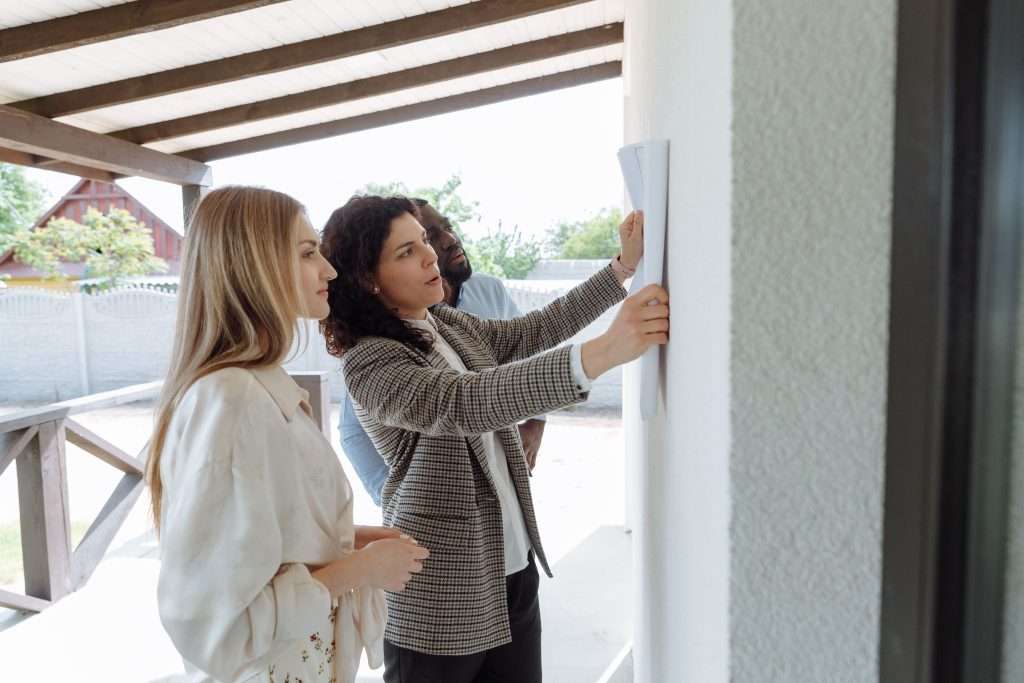 Real estate agent holding architectural plans against an exterior wall while presenting a house to a couple on a covered porch