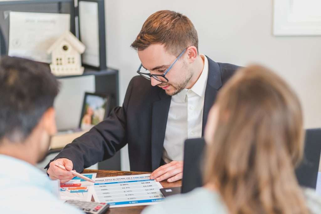 Financial advisor in a suit and glasses pointing at a data chart with a pen while consulting with two clients at an office desk