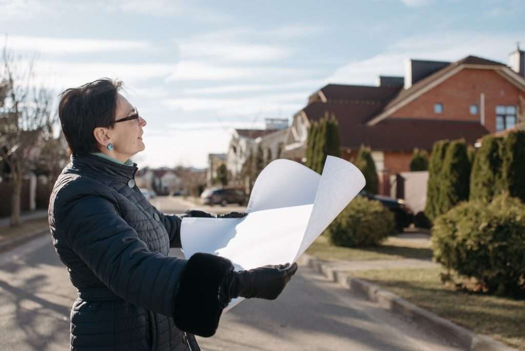 Woman wearing a black puffer coat and glasses reviewing an architectural blueprint outdoors on a residential street