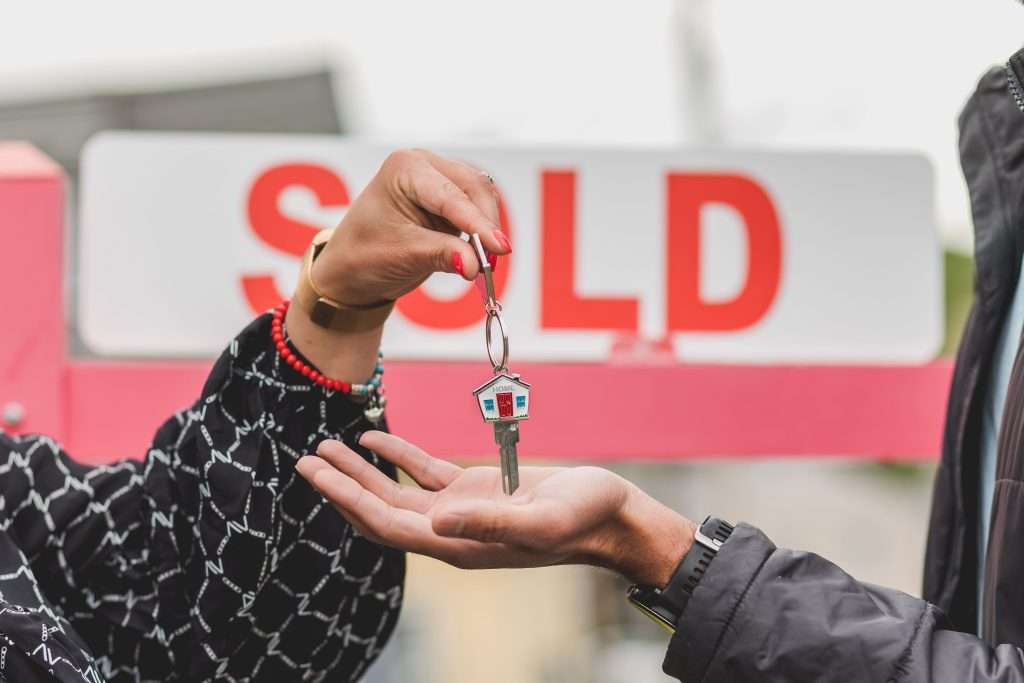 Real estate agent handing over a house key with a small house keychain in front of a red and white "SOLD" sign