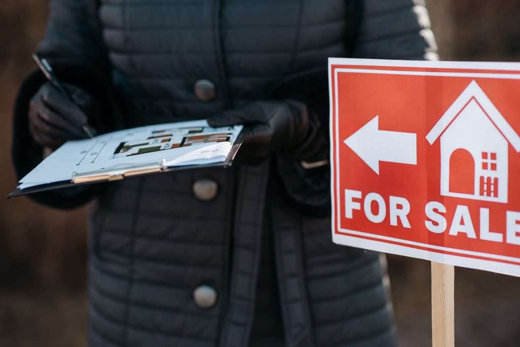 Real estate agent in a black puffer coat holding a clipboard with floor plans next to a red "FOR SALE" sign with an arrow