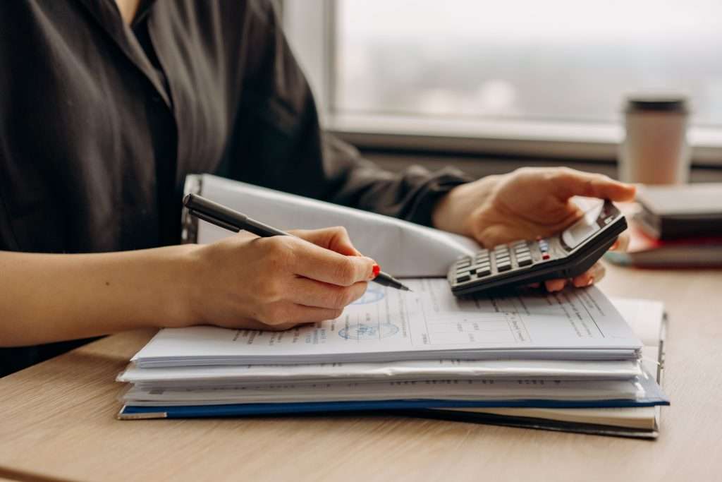 Close-up of a person holding a calculator and pen while reviewing and calculating figures on financial paperwork stacked on a wooden desk