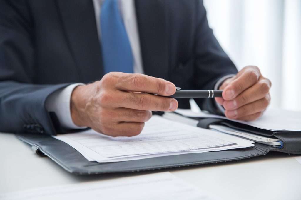 Professional in a dark business suit holding a black pen over signed paperwork inside a leather binder folder on an office desk