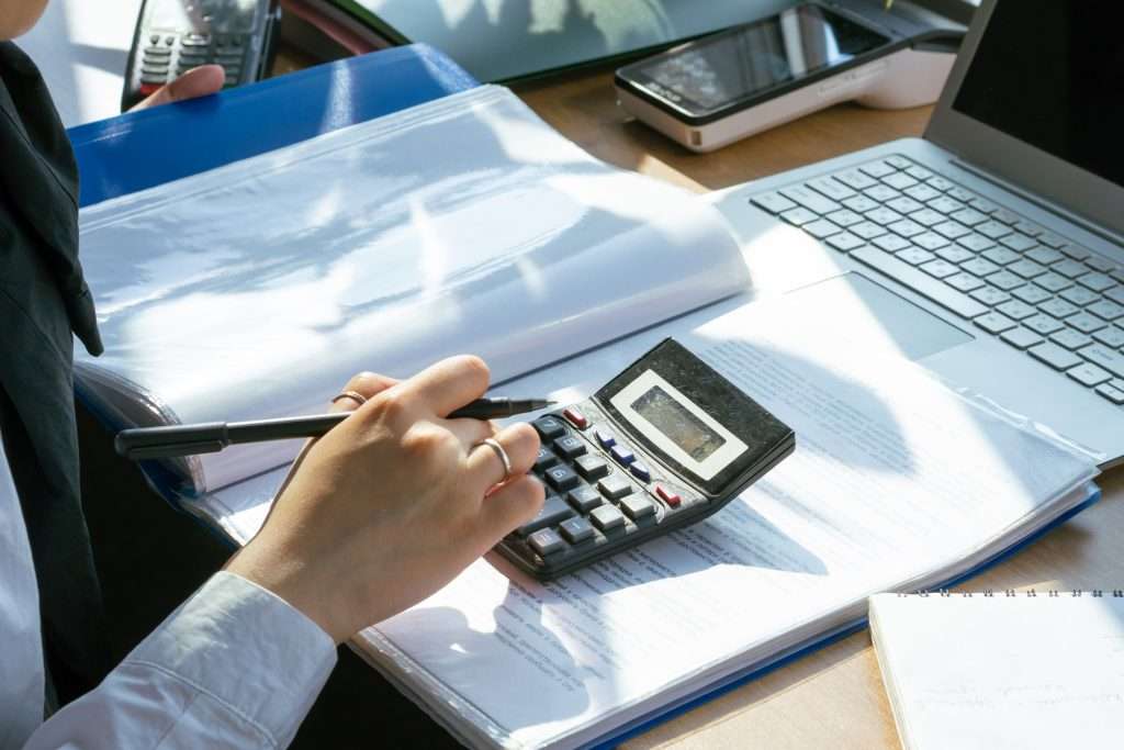 Office professional using a desktop calculator while checking document line items in a binder next to an open laptop